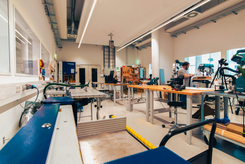 Eine geräumige, helle Werkstatt mit mehreren Werkbänken, Werkzeugen und Geräten. Im Hintergrund arbeitet eine Person an einem Tisch. Große Fenster und eine geordnete Werkzeugaufbewahrung spiegeln die Effizienz der Wirtschaftsinformatik 2.0 wider. at Hochschule Coburg