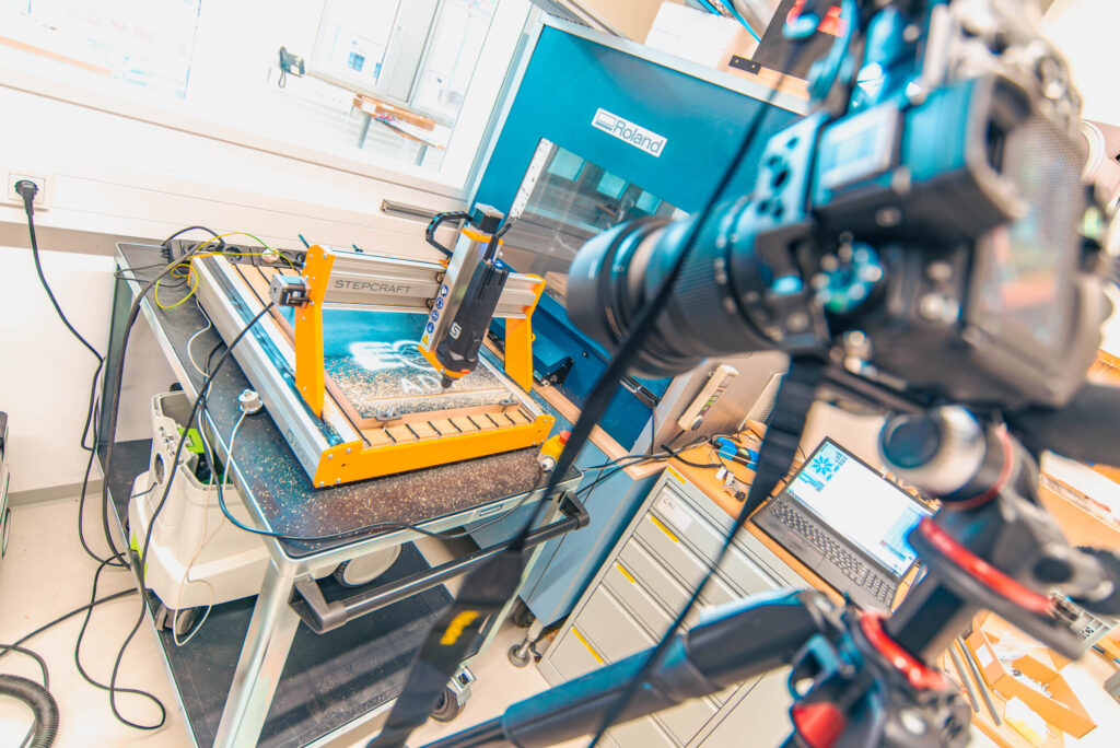 Eine Kamera auf einem Stativ filmt eine kleine CNC-Fräsmaschine in einer hellen Werkstatt, im Hintergrund ein Computer und eine blaue Roland-Maschine – eine Demonstration moderner Produktionsmethoden, inspiriert von der Wirtschaftsinformatik 2.0. at Hochschule Coburg