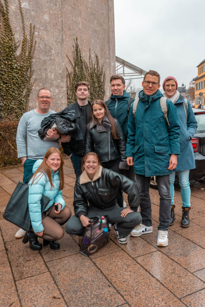 Eine Gruppe von acht jungen Erwachsenen, möglicherweise Studierende der Wirtschaftsinformatik 2.0, posiert und lächelt gemeinsam im Freien auf einem nassen, gefliesten Gehweg mit einem Steingebäude und bunten Häusern im Hintergrund. Die meisten tragen Jacken und Freizeitkleidung. at Hochschule Coburg