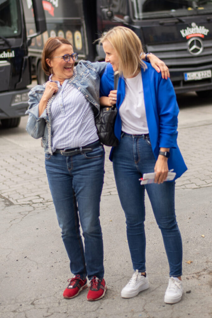 Zwei Frauen stehen dicht beieinander im Freien und haben sich die Arme um die Schultern gelegt. Leger gekleidet, lächeln sie vor schwarzen Trucks mit weißer Schrift – vielleicht nach einer Veranstaltung der Wirtschaftsinformatik. at Hochschule Coburg