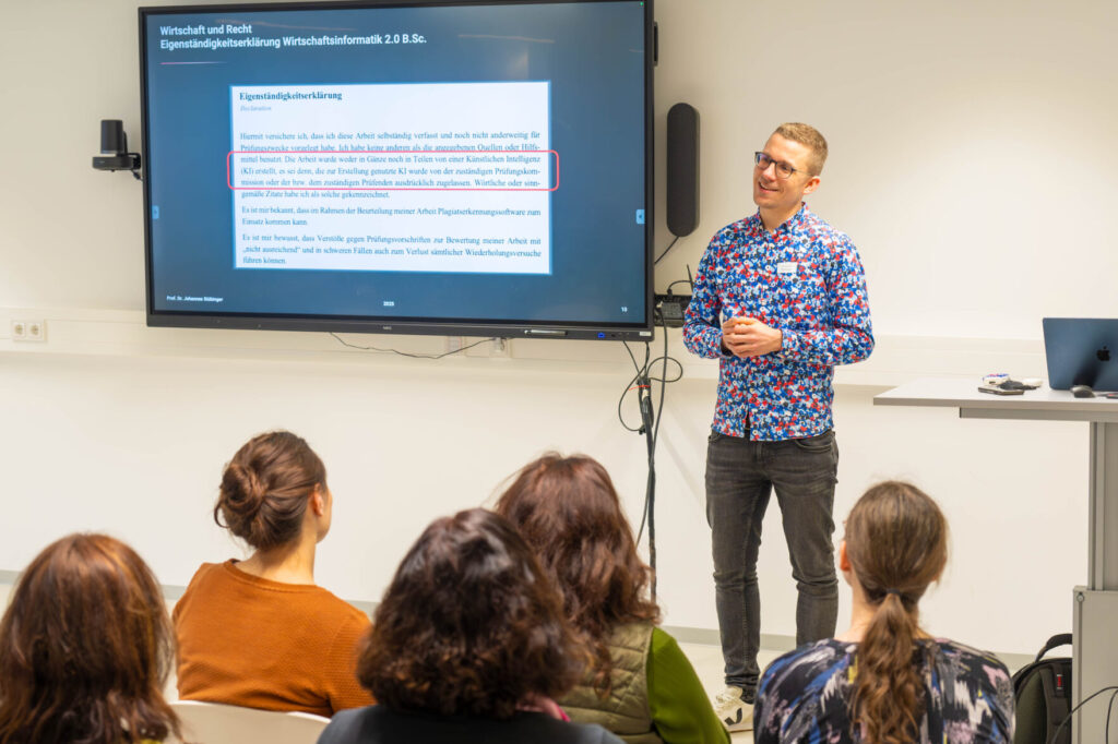 Ein Mann in einem bunten Hemd steht vor einem großen Bildschirm und hält in einem modernen Klassenzimmer eine Präsentation zum Thema Wirtschaftsinformatik 2.0. Auf dem Bildschirm wird deutscher Text angezeigt, während mehrere Personen aufmerksam zuhören. at Hochschule Coburg