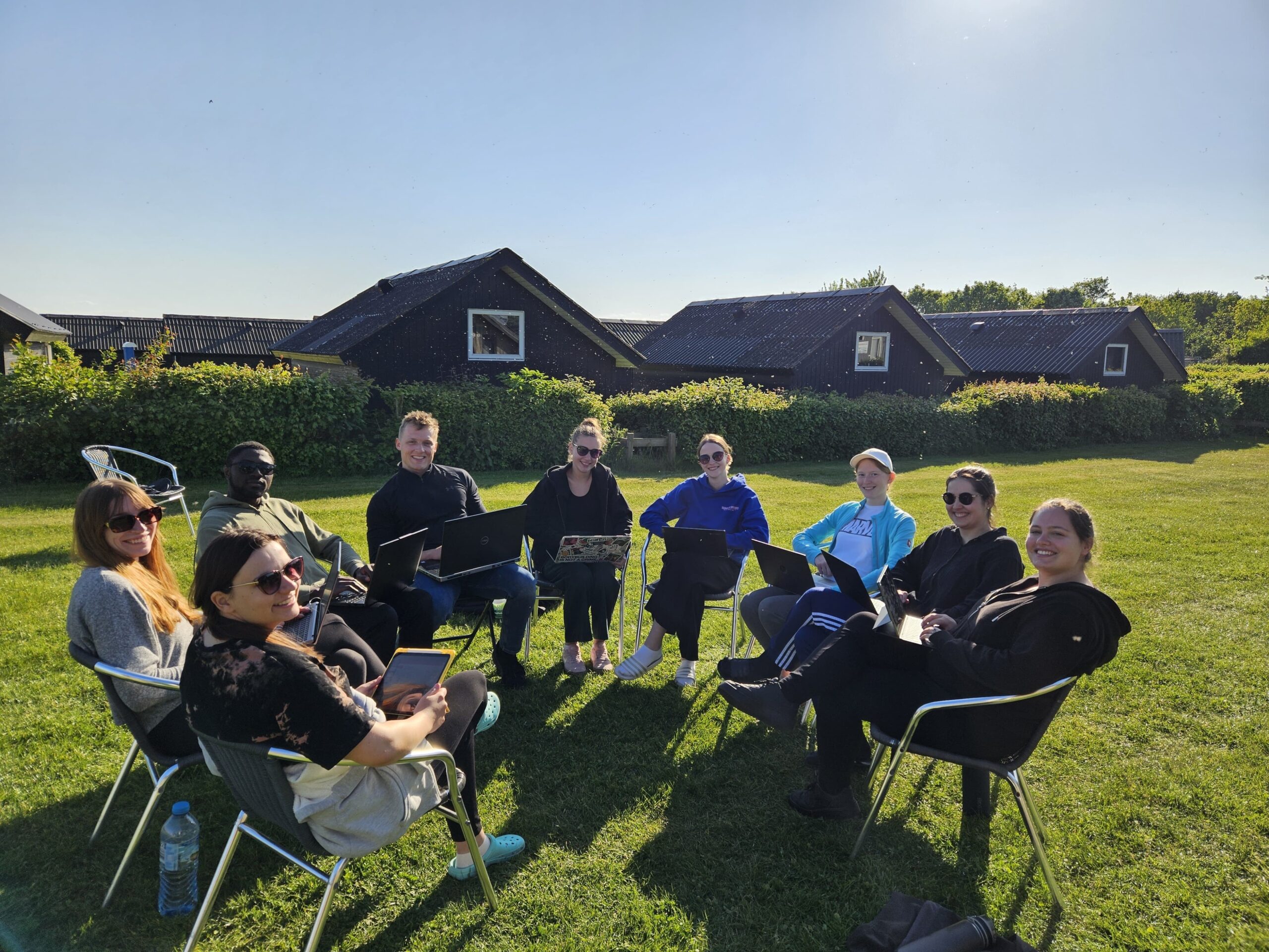 Eine Gruppe von neun Personen sitzt in einem Stuhlkreis auf einer Wiese und diskutiert über Themen wie Bioanalytik und Genreregulierung. Einige haben Laptops und genießen den sonnigen Tag mit Häusern und grünen Büschen im Hintergrund unter einem klaren blauen Himmel. at Hochschule Coburg
