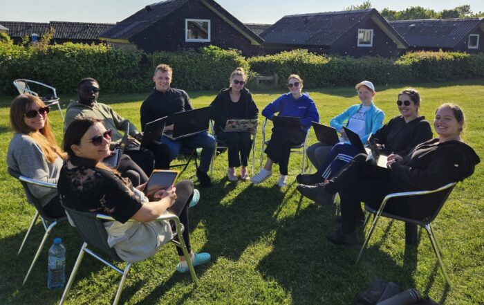 Eine Gruppe von neun Personen sitzt lächelnd im Kreis auf Stühlen im Freien, mit Laptops auf dem Schoß, und diskutiert über Genreregulierung. Sie stehen auf einer grünen Wiese, im Hintergrund sind kleine Häuser und Büsche zu sehen, unter einem strahlend blauen Himmel. at Hochschule Coburg
