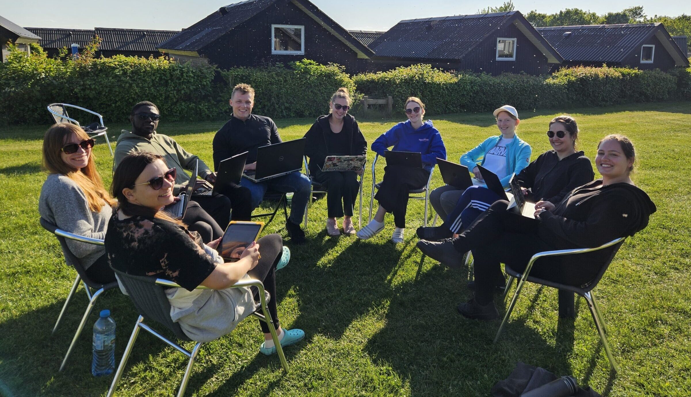 Eine Gruppe von neun Personen sitzt lächelnd im Kreis auf Stühlen im Freien, mit Laptops auf dem Schoß, und diskutiert über Genreregulierung. Sie stehen auf einer grünen Wiese, im Hintergrund sind kleine Häuser und Büsche zu sehen, unter einem strahlend blauen Himmel. at Hochschule Coburg
