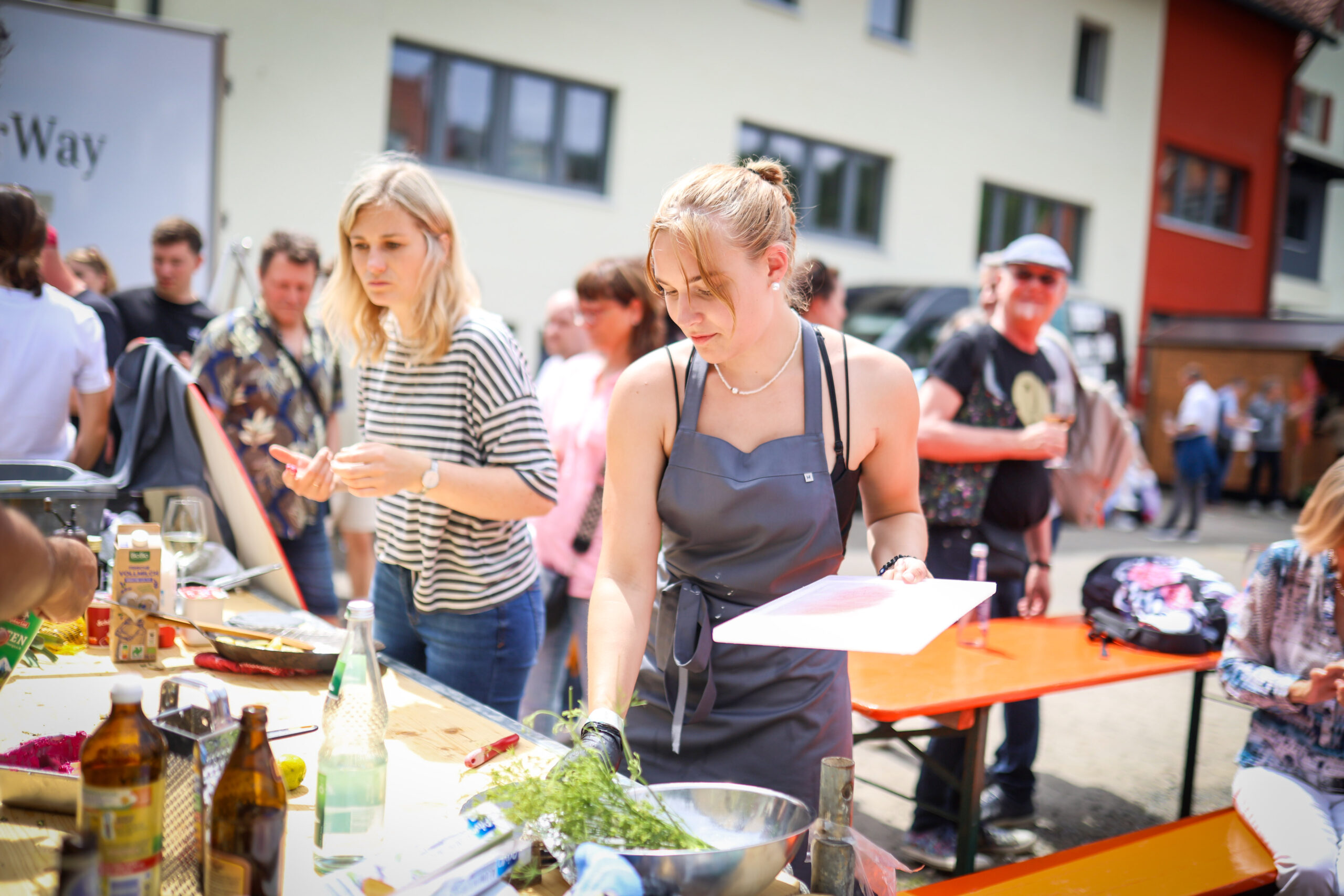 Bei der Hochschul-Grillmeisterschaft bereitet eine Frau in einer grauen Schürze mit einem Schneidebrett in der Hand Essen zu. Die Leute versammeln sich mit Zutaten und Küchenutensilien um Tische und genießen eine sonnige, entspannte Atmosphäre, die Gemeinschaft fördert. at Hochschule Coburg