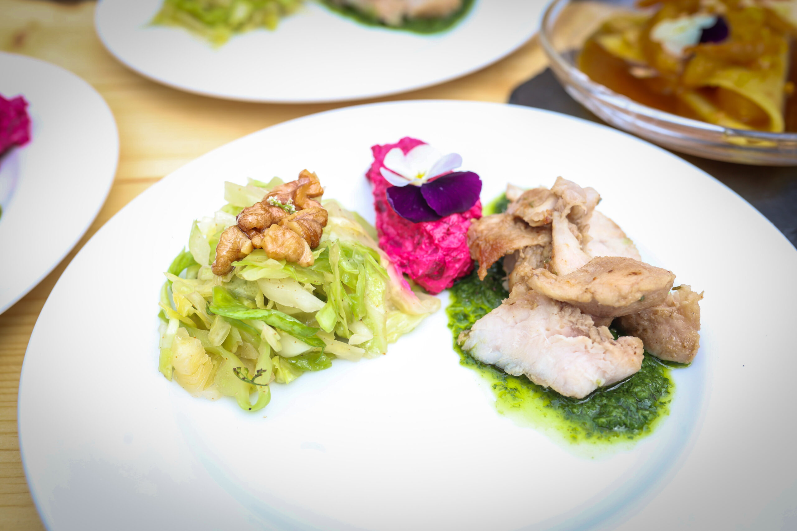 Ein weißer Teller mit sautiertem Kohl, garniert mit Walnüssen, gebratenen Fleischscheiben auf grüner Sauce und einem Klecks rosa Rübenpüree, garniert mit einer essbaren Blüte – Nachhaltigkeit bei der Hochschul-Grillmeisterschaft. Im Hintergrund sind weitere Gerichte zu sehen. at Hochschule Coburg