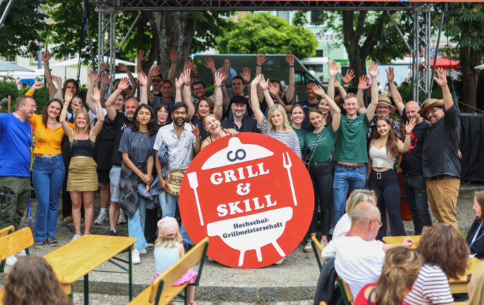 Eine große Gruppe von Menschen posiert und winkt unter einem rosa „ZU TISCH! 25“-Banner bei einer Open-Air-Veranstaltung zur Feier der Gemeinschaft. Davor steht ein Schild mit der Aufschrift „Grill & Skill“ und einer Grillgrafik. Bänke und Bäume runden die festliche Atmosphäre ab. at Hochschule Coburg