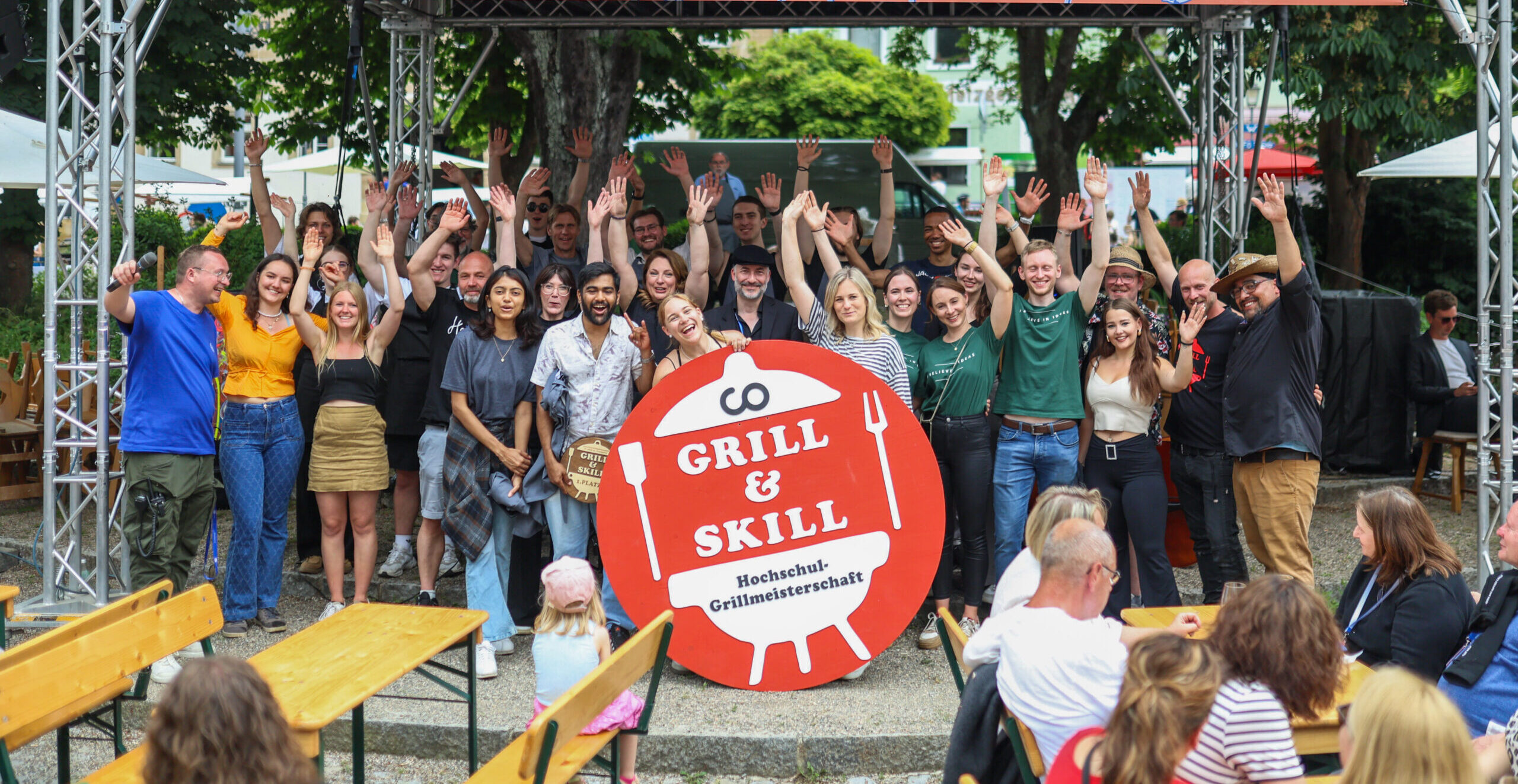 Eine große Gruppe von Menschen posiert und winkt unter einem rosa „ZU TISCH! 25“-Banner bei einer Open-Air-Veranstaltung zur Feier der Gemeinschaft. Davor steht ein Schild mit der Aufschrift „Grill & Skill“ und einer Grillgrafik. Bänke und Bäume runden die festliche Atmosphäre ab. at Hochschule Coburg