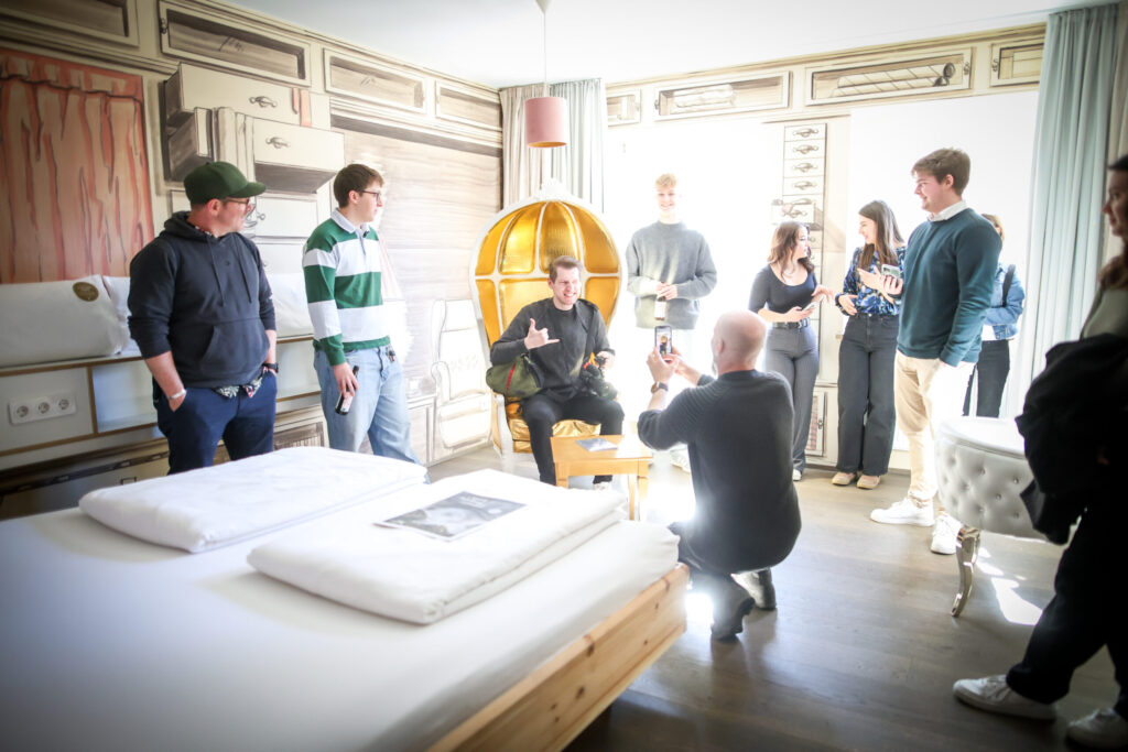 Eine Gruppe von Menschen versammelt sich in einem hell erleuchteten, kunstvoll gestalteten Hotelzimmer. Eine Person sitzt auf einem gelben Stuhl, die anderen stehen da, schauen zu oder machen Fotos. Das einfallende Sonnenlicht schafft eine fröhliche und lebendige Atmosphäre. at Hochschule Coburg