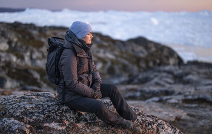 Eine Person in Winterkleidung und mit blauem Hut sitzt auf einem Felsvorsprung und blickt in die Ferne. Im Hintergrund sind schneebedeckte Berge oder Eisberge unter einem sanften Sonnenuntergangshimmel zu sehen. at Hochschule Coburg