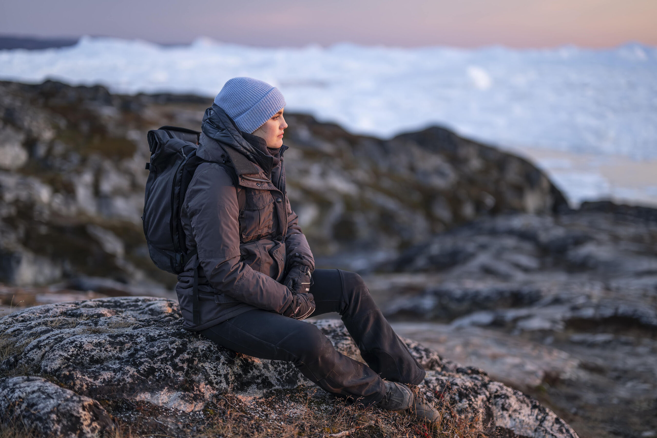 Eine Person in Winterkleidung und mit blauem Hut sitzt auf einem Felsvorsprung und blickt in die Ferne. Im Hintergrund sind schneebedeckte Berge oder Eisberge unter einem sanften Sonnenuntergangshimmel zu sehen. at Hochschule Coburg