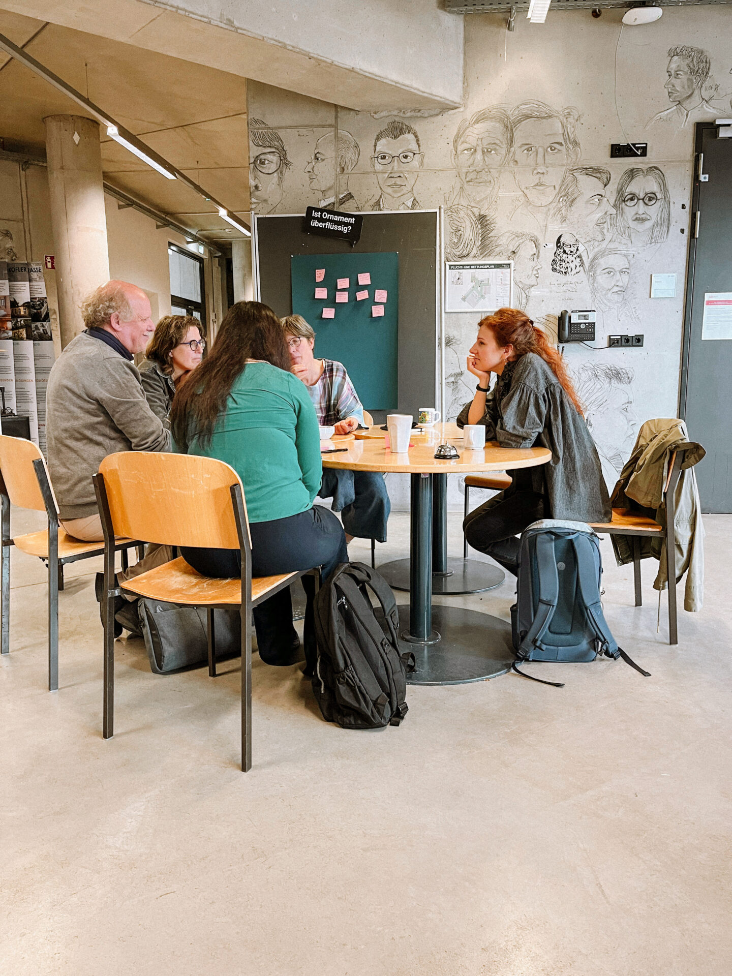 Fünf Personen sitzen in einem modernen Raum um einen kleinen runden Tisch, unterhalten sich und schreiben. Rucksäcke stehen in der Nähe. Dahinter spiegeln eine Tafel mit Haftnotizen und eine Wand mit Porträtskizzen die Zukunftsgestaltung von INSIDE CAMPUS DESIGN für 2025 wider. at Hochschule Coburg