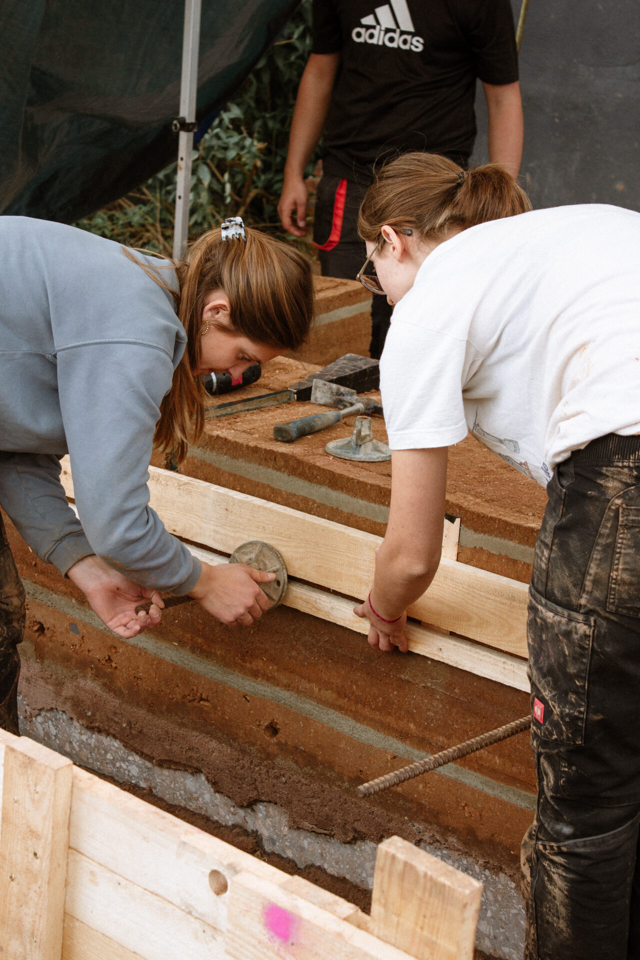 Zwei Personen arbeiten gemeinsam an einem Bauprojekt. Sie glätten und formen Erde oder Beton innerhalb eines Holzrahmens – ein Beispiel für Zukunft gestalten auf dem Campus Design. Eine weitere Person in einem schwarzen Hemd steht daneben und ist teilweise sichtbar. at Hochschule Coburg