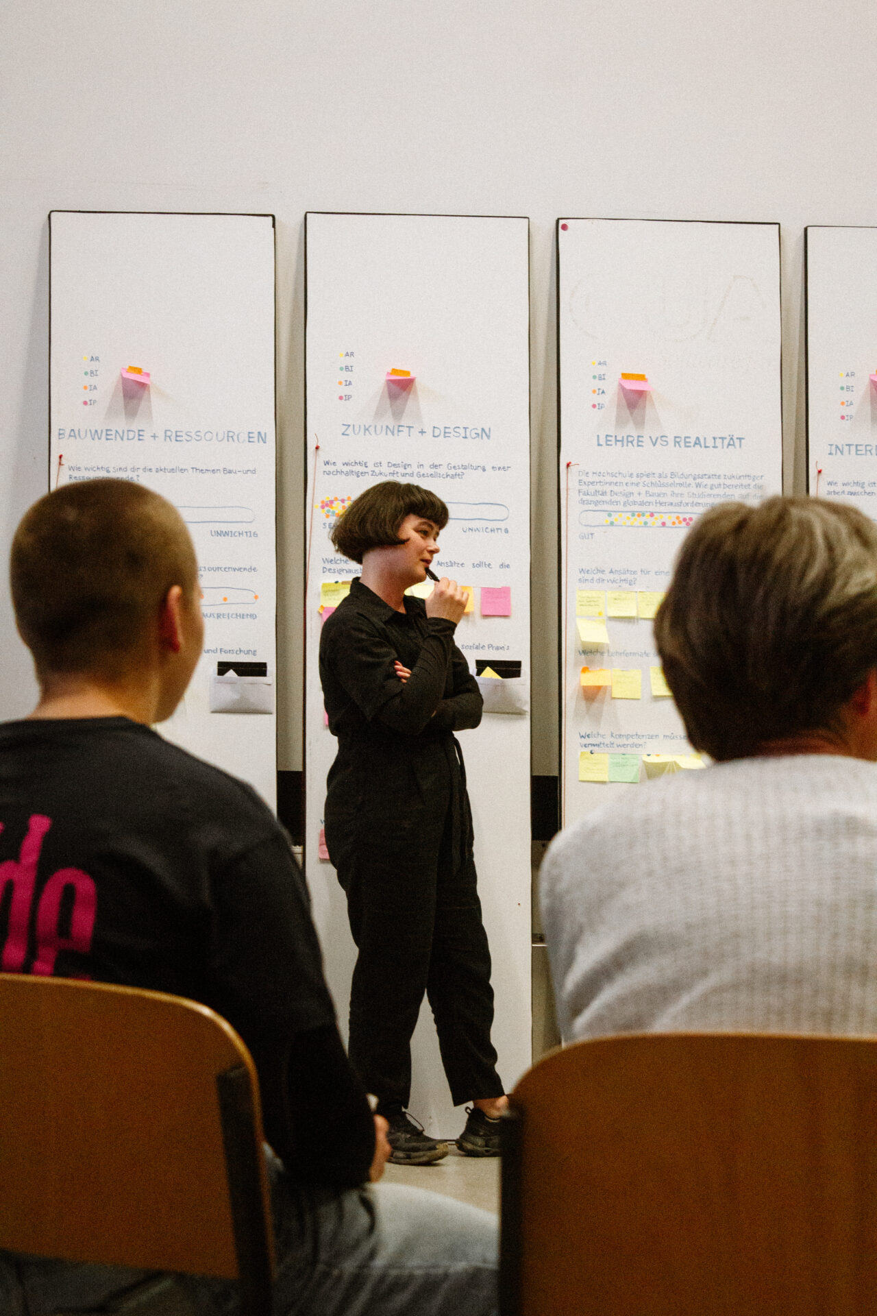 Eine Frau in Schwarz steht und spricht vor drei mit Haftnotizen bedeckten Whiteboards und inspiriert ihr Publikum während eines INSIDE CAMPUS DESIGN-Workshops oder einer Unterrichtsstunde zur Zukunftsgestaltung. at Hochschule Coburg