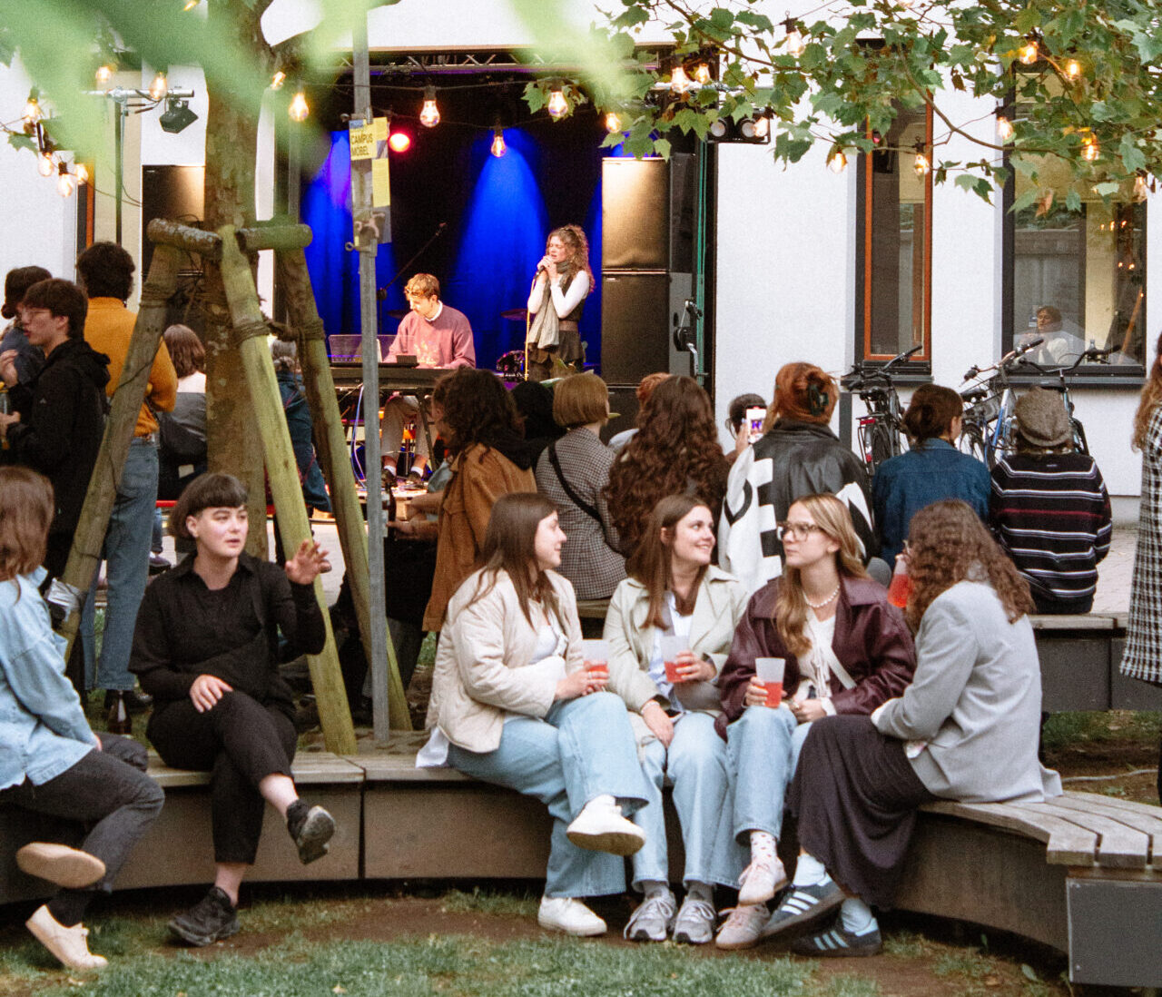 Eine Gruppe von Menschen sitzt und unterhält sich auf geschwungenen Bänken bei einer Veranstaltung von 2025 INSIDE CAMPUS DESIGN im Freien, während eine Band unter blauem Licht auf der Bühne spielt. Lichterketten hängen von Bäumen und schaffen eine festliche Atmosphäre zur Zukunftsgestaltung. at Hochschule Coburg