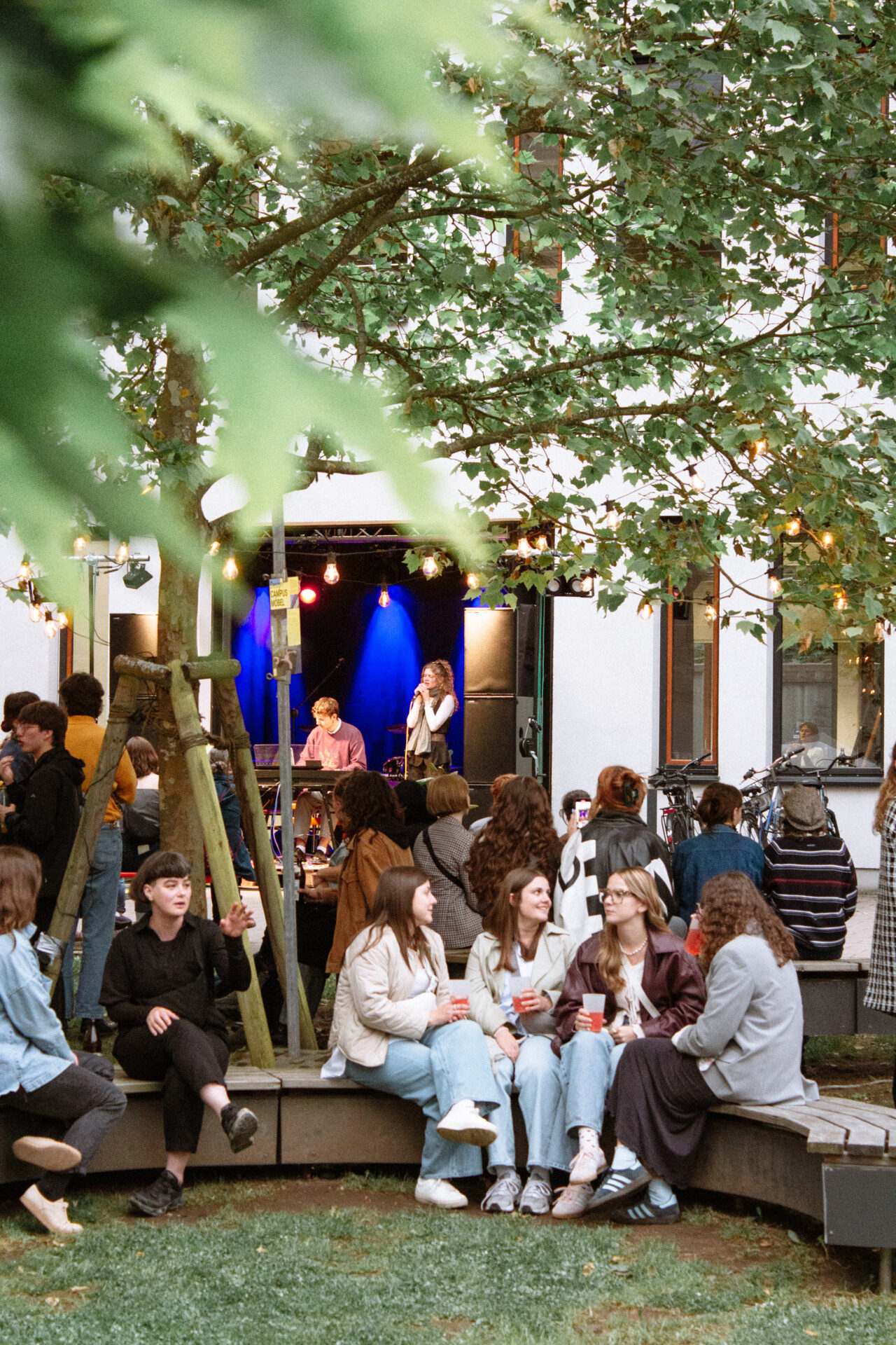 Eine Gruppe von Menschen sitzt und unterhält sich auf geschwungenen Bänken bei einer Veranstaltung von 2025 INSIDE CAMPUS DESIGN im Freien, während eine Band unter blauem Licht auf der Bühne spielt. Lichterketten hängen von Bäumen und schaffen eine festliche Atmosphäre zur Zukunftsgestaltung. at Hochschule Coburg