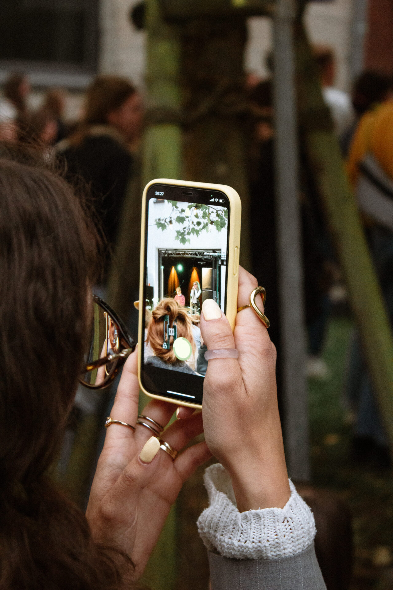 Eine Person mit Ringen und manikürten Nägeln hält ein Smartphone und filmt eine Frau, die draußen für INSIDE CAMPUS DESIGN 2025 posiert. Der Telefonbildschirm zeigt die Szene mit verschwommenen Menschen im Hintergrund – Zukunft gestalten in Aktion. at Hochschule Coburg