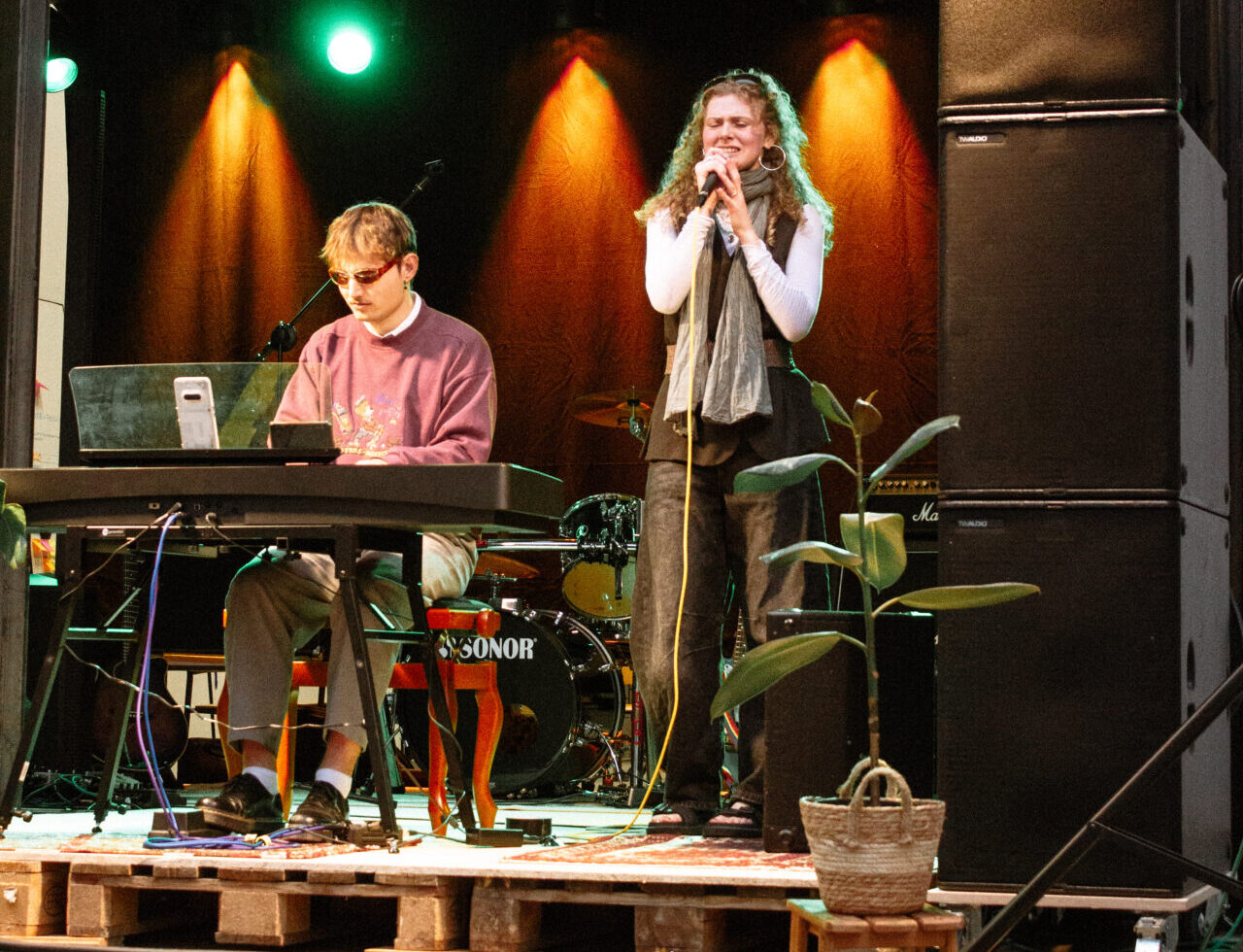 Ein junger Mann spielt Keyboard, während eine junge Frau auf einer kleinen Indoor-Bühne mit grünem Licht in ein Mikrofon singt, umgeben von Musikgeräten und Topfpflanzen – was die kreative Atmosphäre von INSIDE CAMPUS DESIGN 2025 widerspiegelt. at Hochschule Coburg
