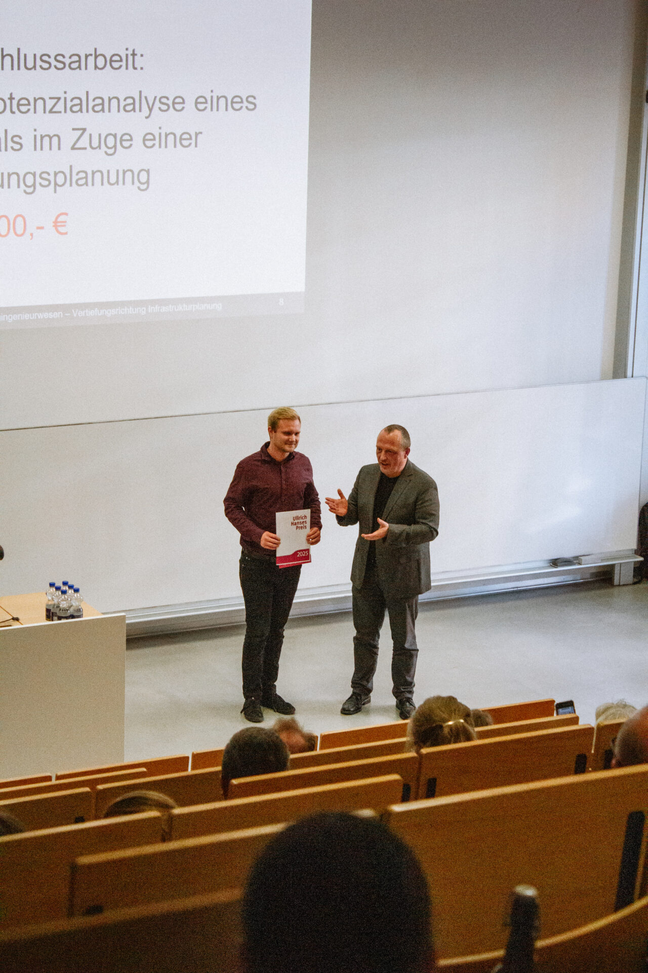 Zwei Männer stehen vor einem Hörsaal. Einer von ihnen hält lächelnd eine Urkunde in der Hand und wird für seine Arbeit an INSIDE CAMPUS DESIGN 2025 ausgezeichnet. Hinter ihnen wird eine Präsentationsfolie von Zukunft gestalten projiziert, während das Publikum zusieht. at Hochschule Coburg
