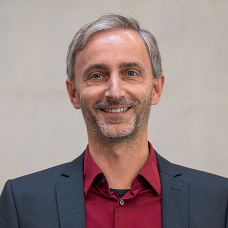 A man with short gray hair and a beard is smiling. He is wearing a dark suit jacket over a burgundy dress shirt and standing in front of a plain, light-colored background. at Hochschule Coburg