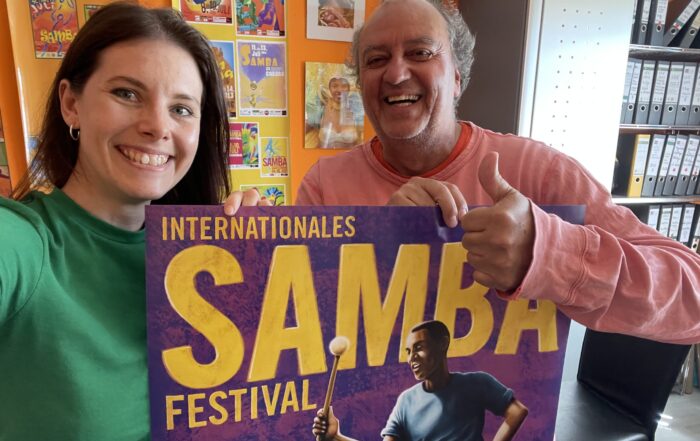 Zwei lächelnde Menschen halten ein buntes "Internationales Samba Festival"-Plakat für den Plakatwettbewerb. Die Frau im grünen Hemd steht links, der Mann in Rosa rechts zeigt den Daumen nach oben. Leuchtende Plakate schmücken die orangefarbene Wand hinter ihnen. at Hochschule Coburg