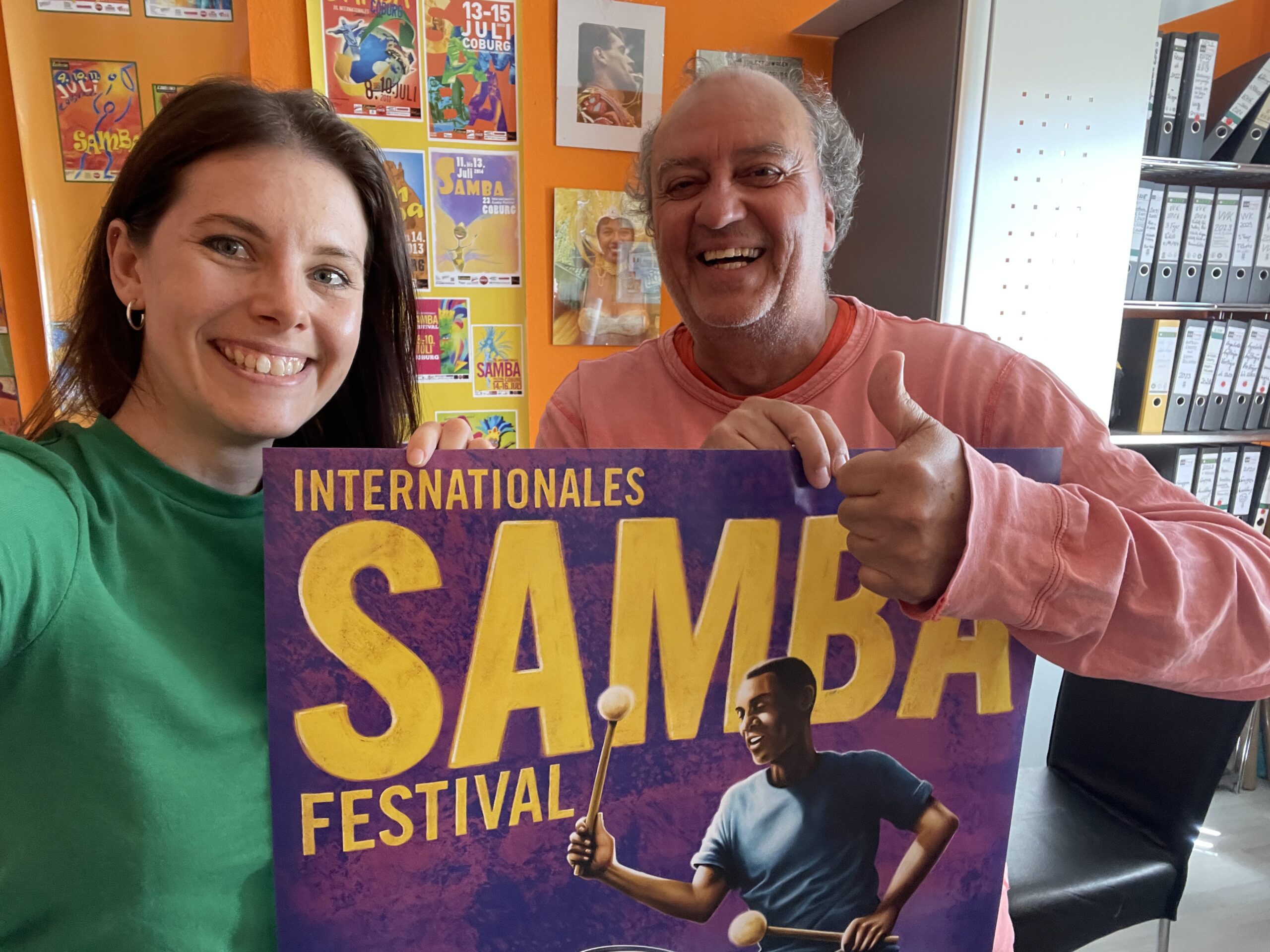 Zwei lächelnde Menschen halten ein buntes "Internationales Samba Festival"-Plakat für den Plakatwettbewerb. Die Frau im grünen Hemd steht links, der Mann in Rosa rechts zeigt den Daumen nach oben. Leuchtende Plakate schmücken die orangefarbene Wand hinter ihnen. at Hochschule Coburg