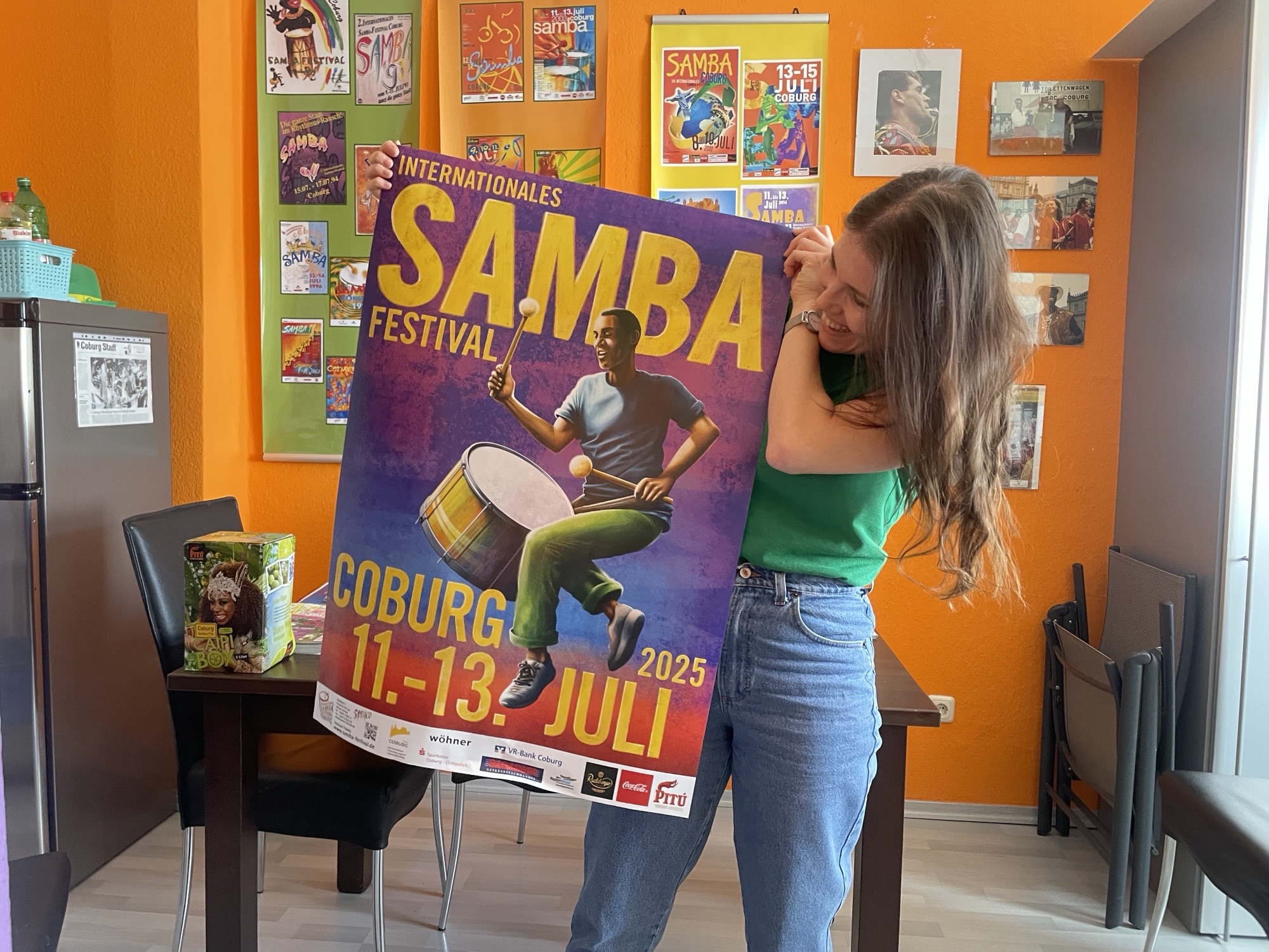 Eine lächelnde Frau, Vanessa Gundler, hält ein buntes Plakatwettbewerb-Plakat für das Samba-Festival 2025 in Coburg, auf dem ein Schlagzeuger zu sehen ist. Zeitschriftencover und Samba-Plakate schmücken die leuchtend orangefarbene Wand hinter ihr. at Hochschule Coburg