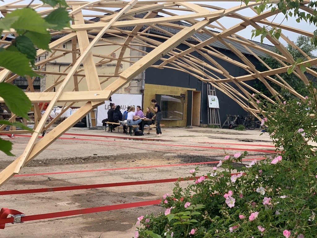 Mehrere Personen sitzen an einem Tisch unter einem teilweise aufgebauten Holzrahmen, umgeben von Baumaterialien. Rosa Blumen und grüne Blätter stehen im Vordergrund, während das große Gebäude auf innovatives CAMPUS DESIGN hindeutet, das die Zukunftsgestaltung von 2025 prägt. at Hochschule Coburg