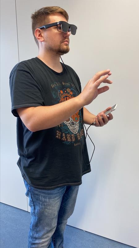Ein Mann mit einer Datenbrille steht in einem Gebäude, hält in einer Hand ein kleines, durch ein Kabel verbundenes Gerät und hebt die andere Hand mit leicht gekrümmten Fingern an, als würde er AR-Technologie verwenden oder die Möglichkeiten des Visual Computing erkunden. at Hochschule Coburg