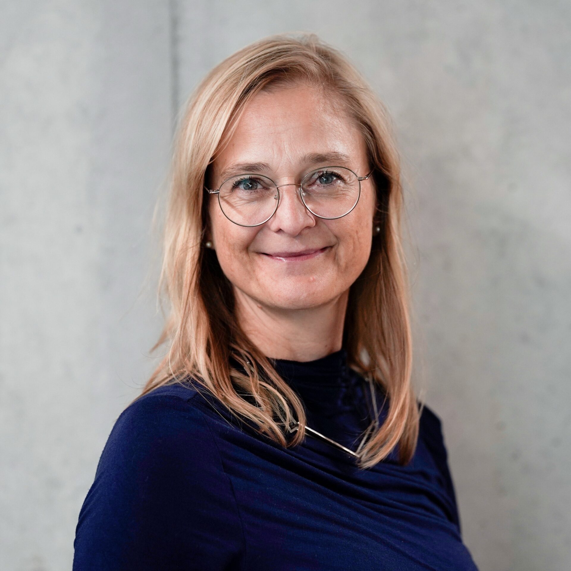 A woman with long blonde hair and glasses smiles at the camera. She is wearing a navy blue top and a silver necklace, standing against a plain light gray background. at Hochschule Coburg