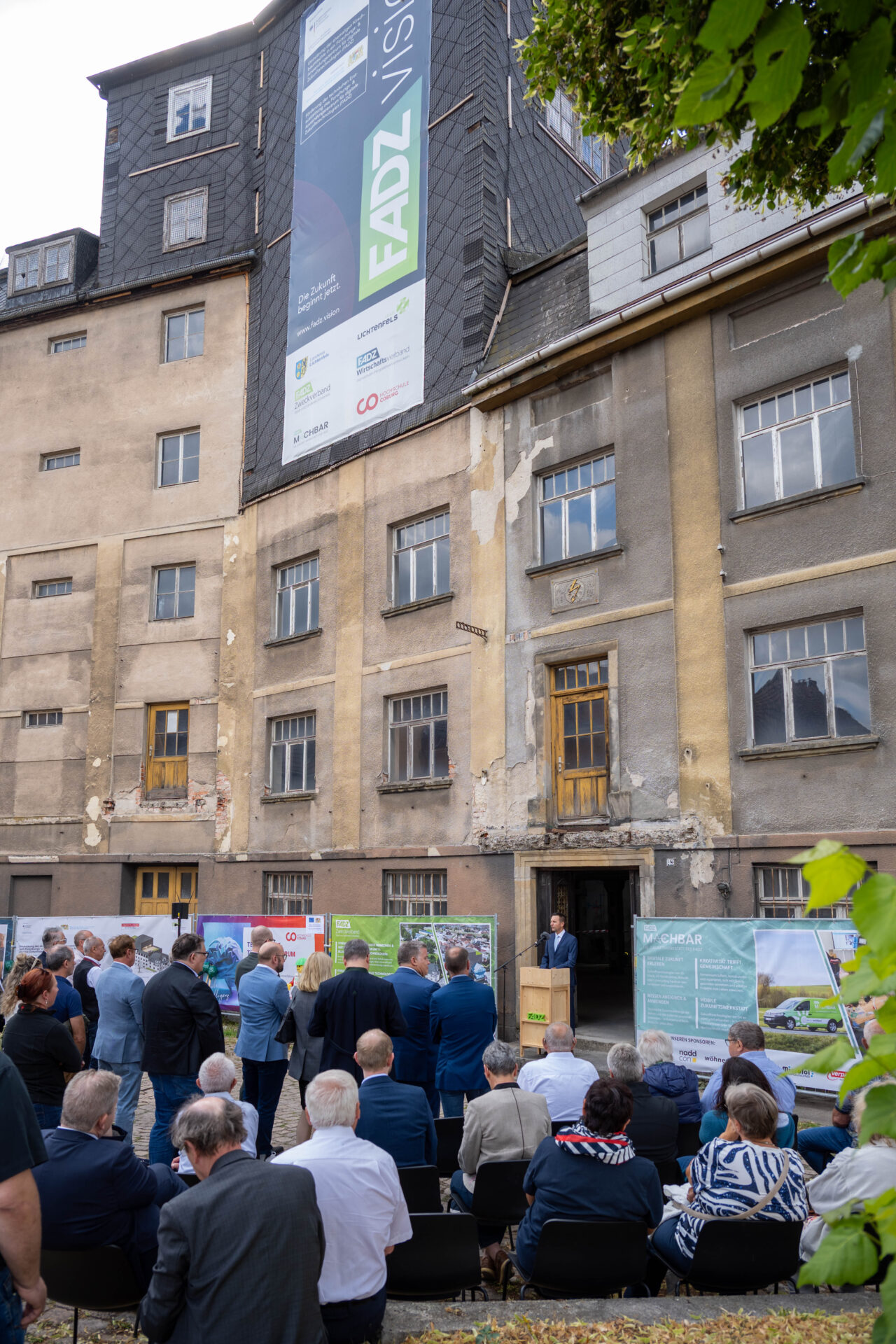 Vor der verwitterten Kirschbaummühle, deren Fassade mit Bannern geschmückt ist, versammelt sich eine Gruppe von Menschen, die an einem sonnigen Tag an einer Freiluftveranstaltung zur digitalen Zukunft teilnehmen. at Hochschule Coburg