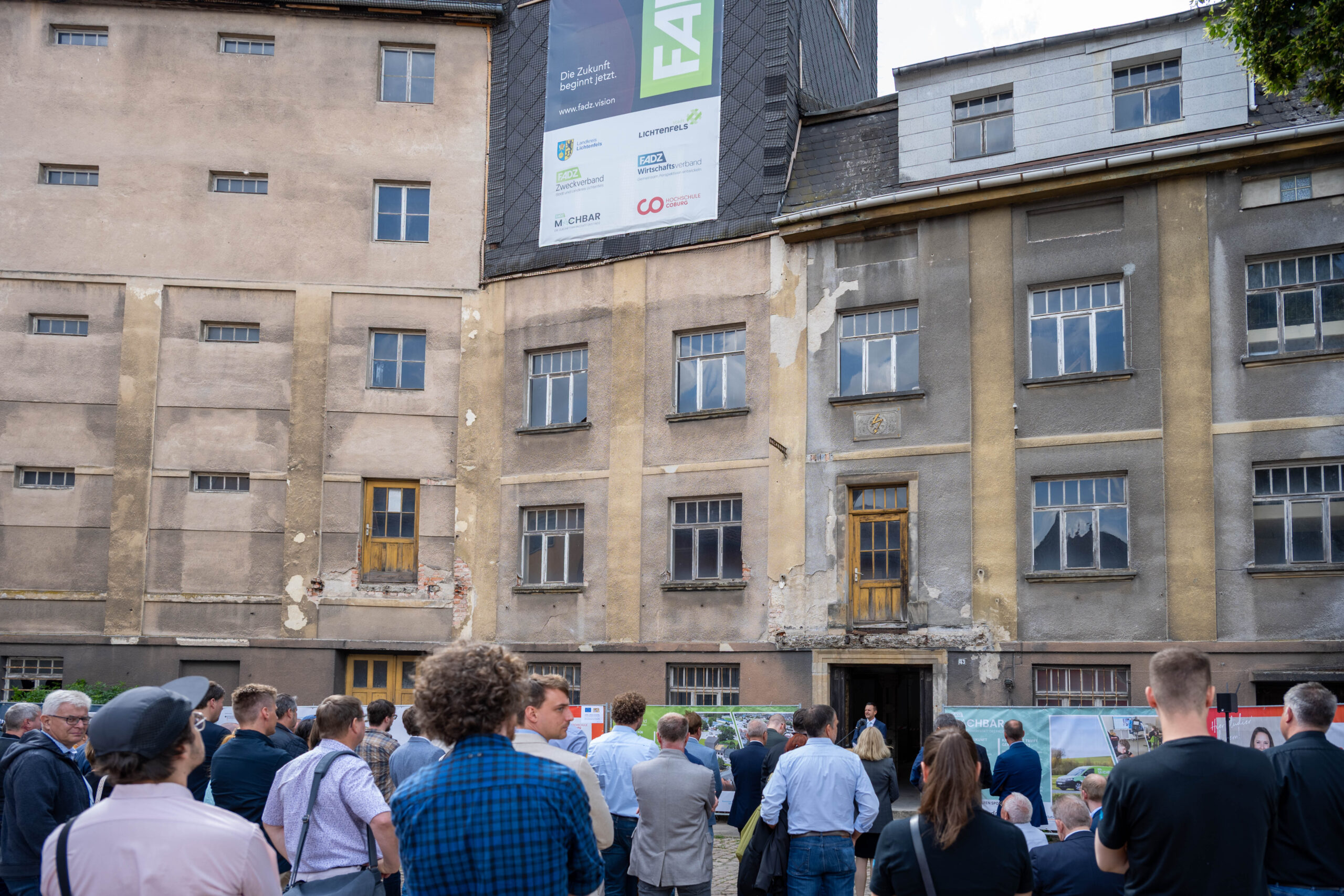 Vor der verwitterten Kirschbaummühle mit ihrer abblätternden Farbe und den vielen Fenstern, die ein großes Transparent einrahmen, versammelt sich eine Menschenmenge. Einige betrachten Schautafeln, die den Spatenstich für eine digitale Zukunft markieren. at Hochschule Coburg