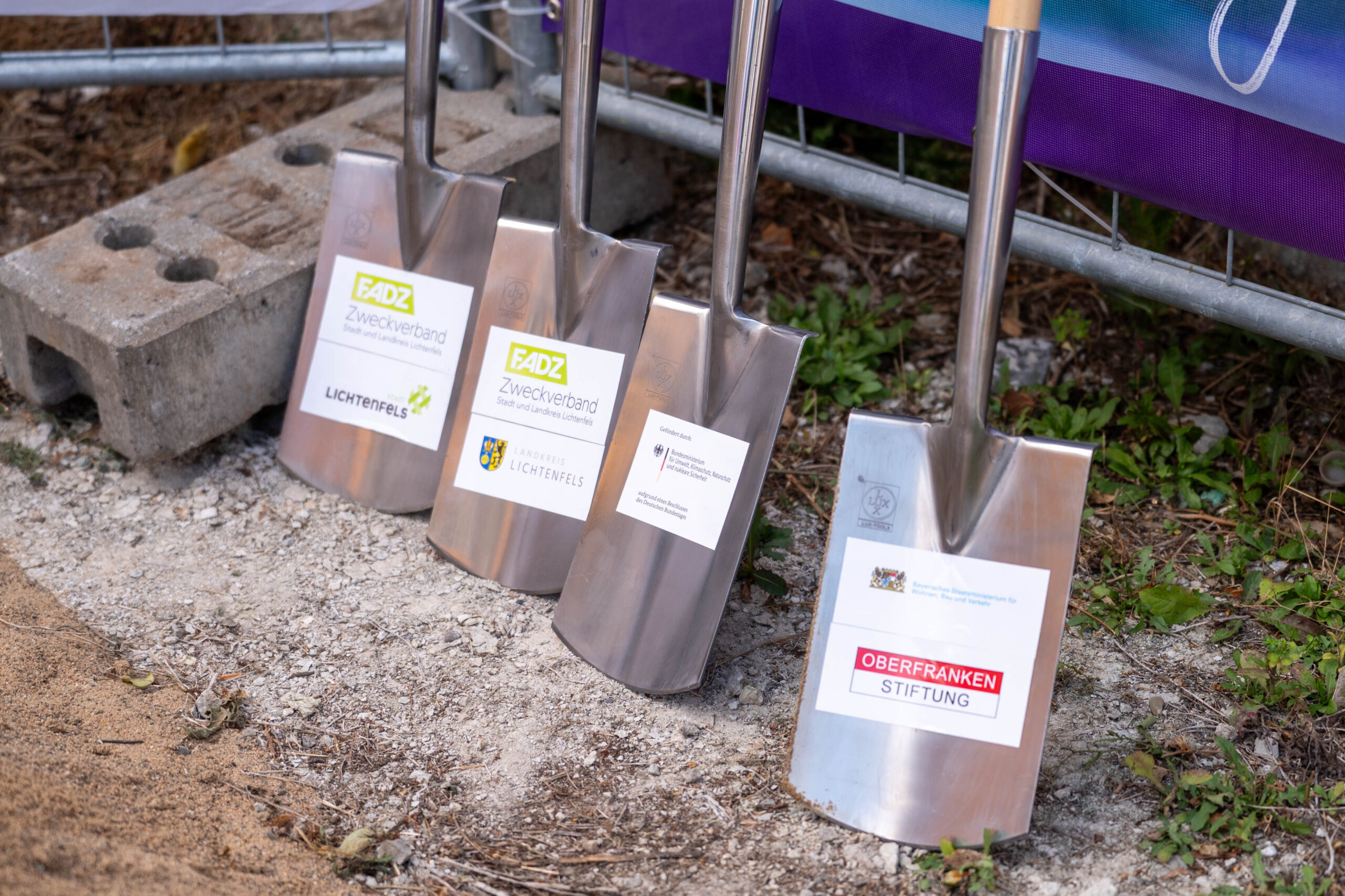 Vier glänzende Schaufeln mit Etiketten stehen an einem Betonklotz und einem Metallzaun auf Schotter und Erde, bereit für den Spatenstich an der Lichtenfelser Kirschbaummühle - ein neuer Schritt zur Gestaltung unserer digitalen Zukunft. at Hochschule Coburg