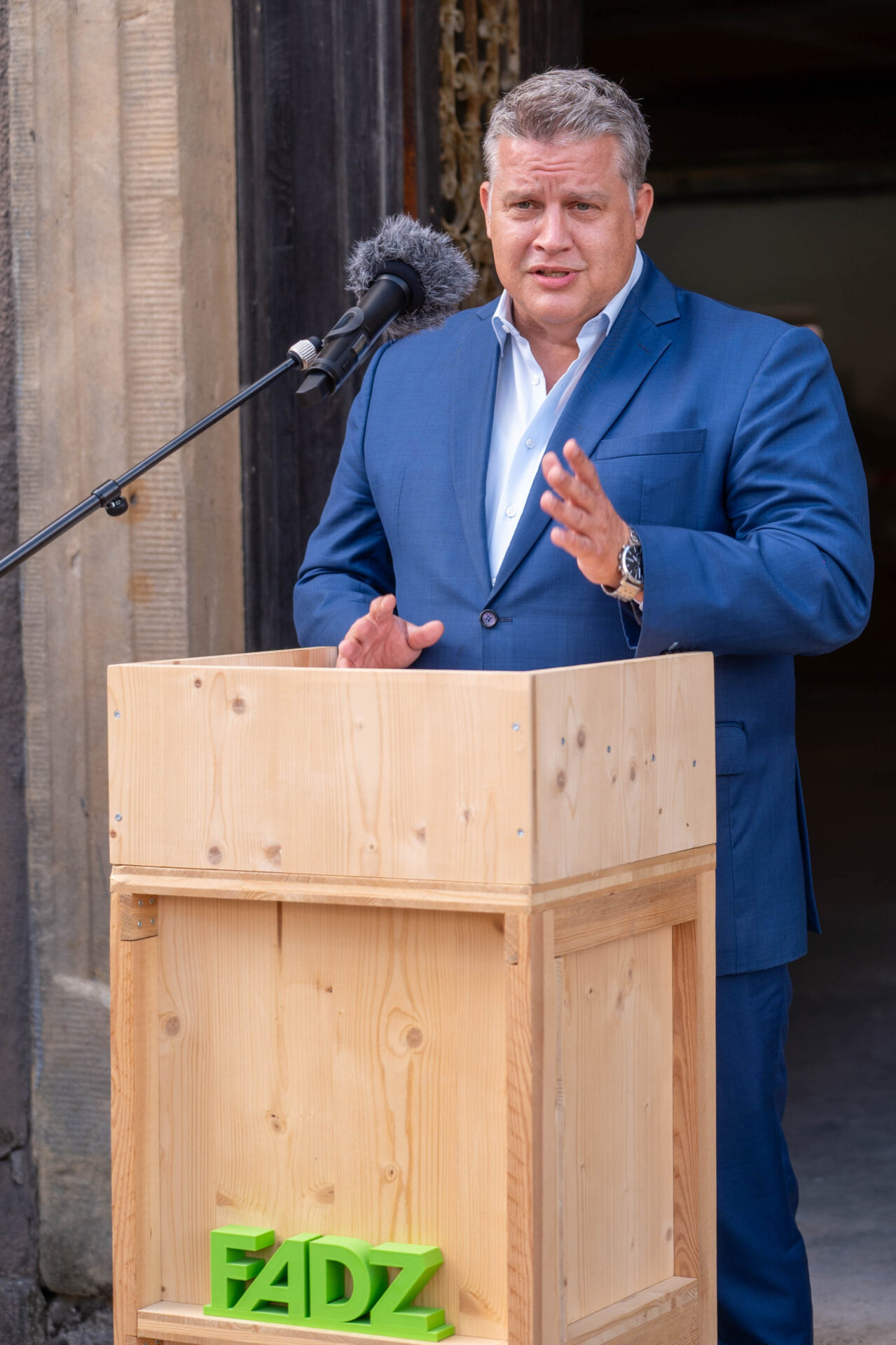 Ein Mann im blauen Anzug spricht an einem hölzernen Podium mit grünem FADZ-Logo und deutet mit Gesten auf den Spatenstich an der Kirschbaummühle und die digitale Zukunft hin, während er vor einem steinernen Tor steht. at Hochschule Coburg