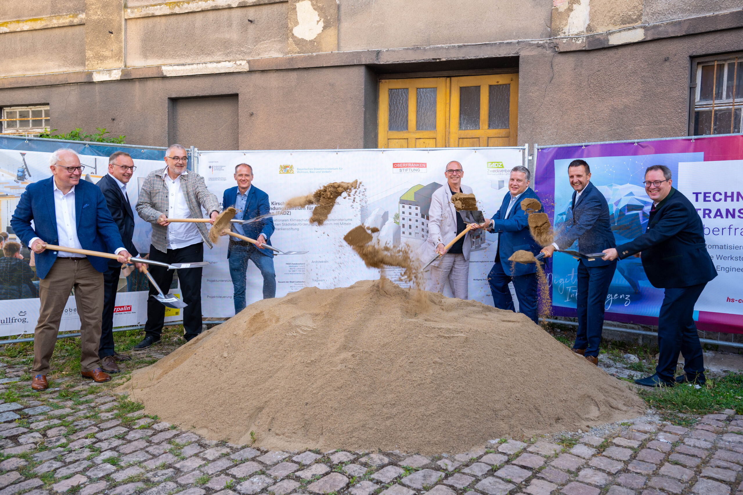 Neun Männer in Geschäftskleidung nehmen an der Spatenstich-Zeremonie an der Kirschbaummühle teil. Sie werfen Sand mit Schaufeln vor einer Baustelle, die den Beginn einer digitalen Zukunft markiert, mit Bannern und einem alten Gebäude im Hintergrund. at Hochschule Coburg