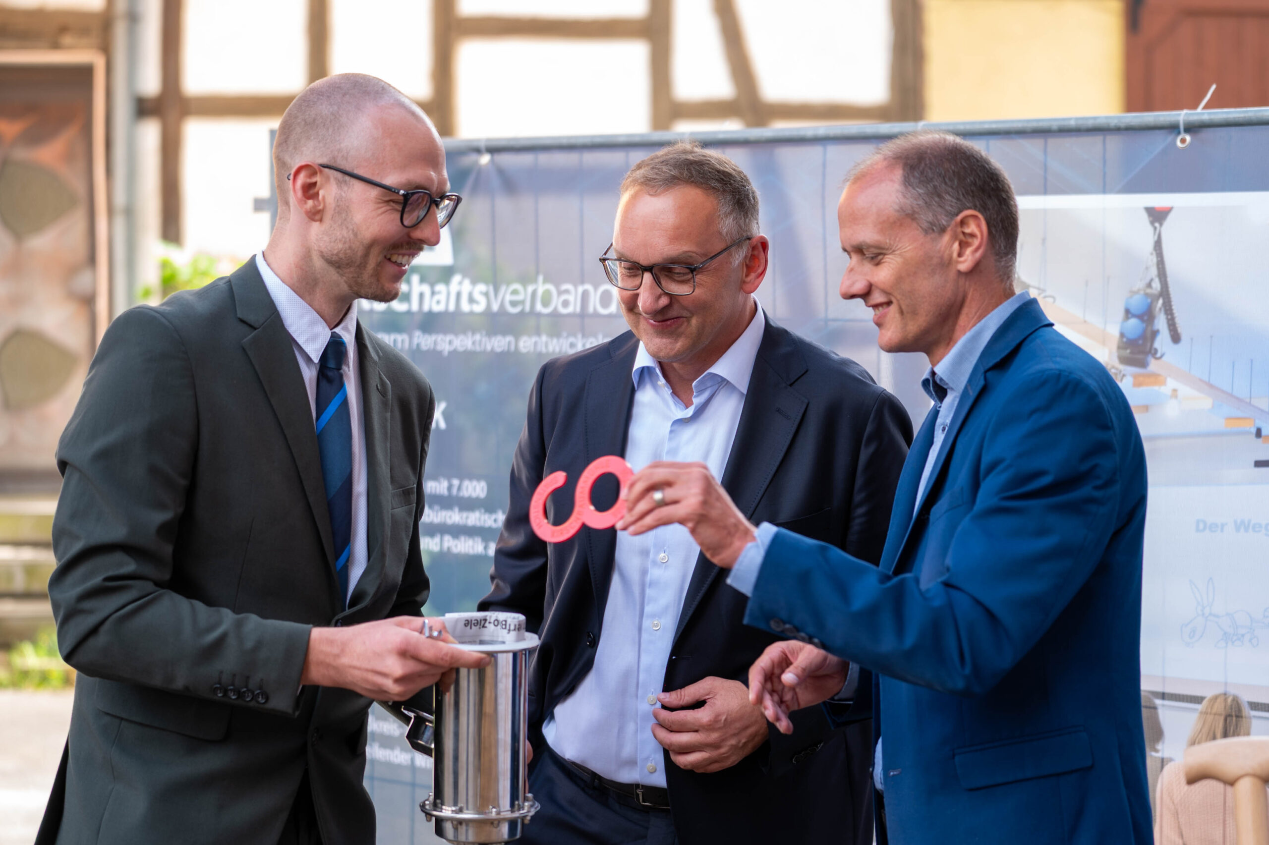 Drei Männer in Anzügen stehen draußen an der Lichtenfelser Kirschbaummühle Spatenstich und lächeln, während sie mit einem Metallbehälter und einem roten Unendlichkeitssymbol interagieren. Im Hintergrund sind ein Banner und ein Gebäude zu sehen, die Schritte in Richtung einer digitalen Zukunft markieren. at Hochschule Coburg