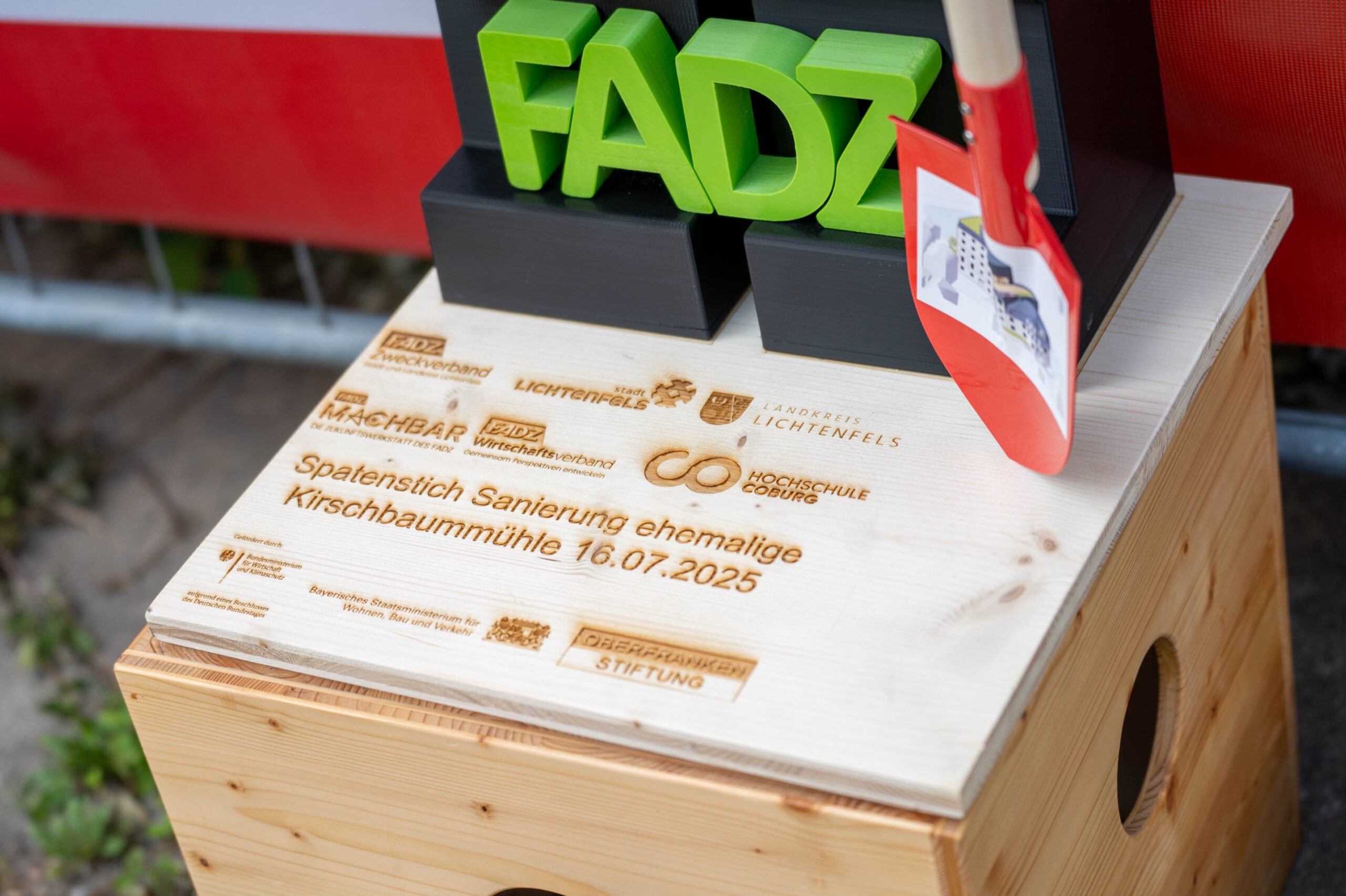 Nahaufnahme eines Holzkastens mit eingraviertem Text und Logos, einem grünen "FADZ"-Schild und einer roten Schaufel, die auf der Oberseite ruht und den Spatenstich für die digitale Zukunft der Lichtenfelser Kirschbaummühle einfängt. Deutlich sichtbar ist das Veranstaltungsdatum "16.07.2025". at Hochschule Coburg