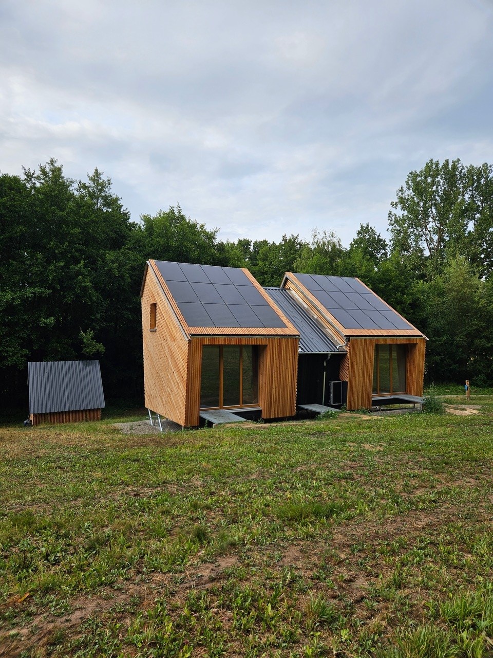 Zwei kleine moderne Holzhäuser mit großen Solarzellen auf den Dächern stehen auf einer Wiese in der Nähe eines Waldgebiets und präsentieren ein nachhaltiges Modulsystem; in der Nähe steht ein kleineres Holzgebäude. Der Himmel ist bedeckt. at Hochschule Coburg