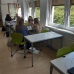 In einem hellen, modern eingerichteten Klassenzimmer sitzt eine Gruppe von Schülern an den Tischen und spricht und arbeitet zusammen. Große Fenster lassen natürliches Licht herein, grüne Stühle umgeben die Tische, und einige Rucksäcke liegen auf dem Boden. at Hochschule Coburg