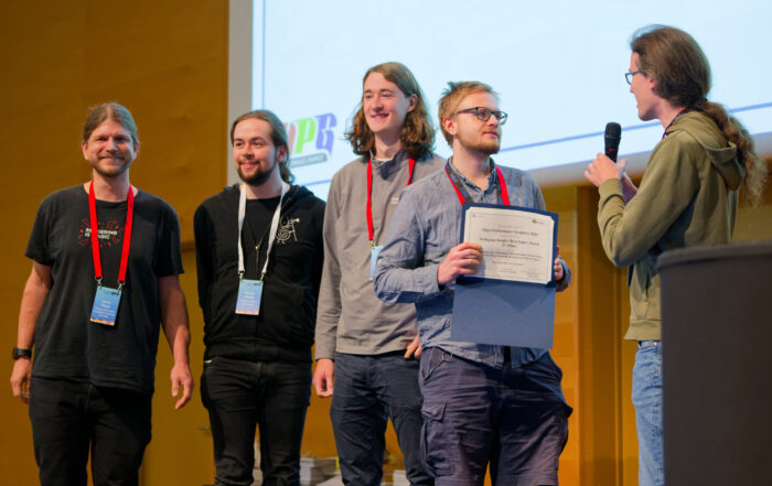 Vier lächelnde Wissenschaftler stehen auf der Bühne; einer hält eine Urkunde, ein anderer spricht mit einem Mikrofon zu ihnen. Eine große Leinwand weist auf den Wissenschaftspreis in Visual-Computing hin. Alle tragen Veranstaltungsplaketten. at Hochschule Coburg
