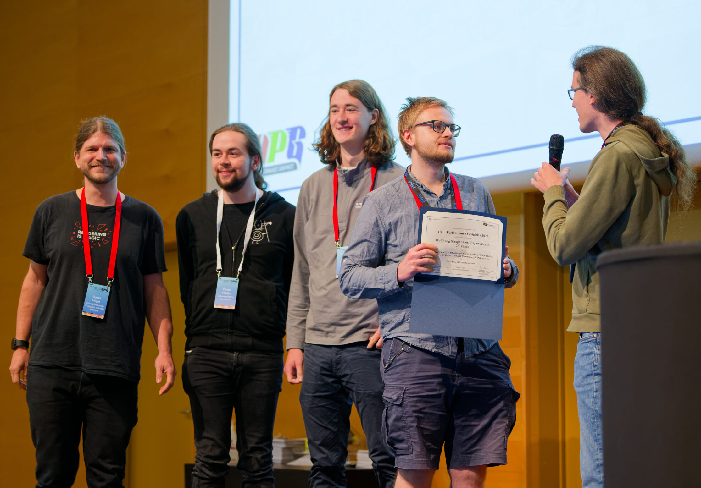 Vier lächelnde Wissenschaftler stehen auf der Bühne; einer hält eine Urkunde, ein anderer spricht mit einem Mikrofon zu ihnen. Eine große Leinwand weist auf den Wissenschaftspreis in Visual-Computing hin. Alle tragen Veranstaltungsplaketten. at Hochschule Coburg