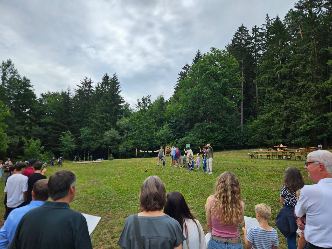 Eine Gruppe von Menschen steht auf einer von Bäumen umgebenen Wiese im Waldkindergarten Pinzberg. Einige Erwachsene und Kinder, die Waldwichtel, versammeln sich in der Mitte, während andere vom Vordergrund aus zusehen. Auf der rechten Seite sind Tische und Bänke aufgestellt. at Hochschule Coburg