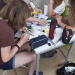 Vier junge Leute sitzen um einen Tisch herum und arbeiten an Bastelprojekten, wobei sie sich von Papier, Schere und Klebstoff inspirieren lassen. Schulsachen und Taschen liegen verstreut in einem scheinbar kreativen Klassenzimmer herum. at Hochschule Coburg