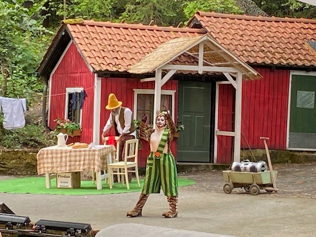 Eine Person im Katzenkostüm posiert spielerisch vor einem roten Holzhäuschen auf der Waldbühne Heldritt, während eine andere mit einem gelben Hut an einem Tisch mit kariertem Tuch steht. In der Nähe hängt Wäsche und auf einem Wagen stehen Milchkannen, die die Märchenzauber-Atmosphäre noch verstärken. at Hochschule Coburg