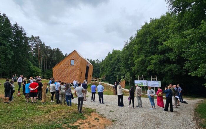 Eine Gruppe versammelt sich auf einem Kiesweg und einer Rasenfläche neben dem modernen, kantigen Waldkindergarten Pinzberg, umgeben von Bäumen unter einem bewölkten Himmel. Schilder weisen auf das nachhaltige Modulsystem in der Nähe des Gebäudes hin. at Hochschule Coburg