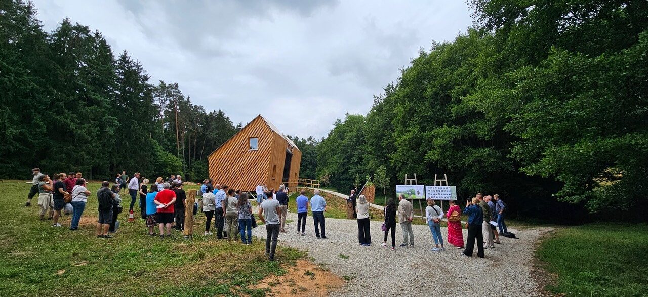 Eine Gruppe versammelt sich auf einem Kiesweg und einer Rasenfläche neben dem modernen, kantigen Waldkindergarten Pinzberg, umgeben von Bäumen unter einem bewölkten Himmel. Schilder weisen auf das nachhaltige Modulsystem in der Nähe des Gebäudes hin. at Hochschule Coburg