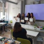 Vier Personen stehen vorne in einem Klassenzimmer und halten Urkunden und von Design inspirierte Kunstwerke in der Hand, während eine andere Person ihnen gegenüber sitzt und ein Foto macht. Der Raum ist mit Tischen, Stühlen und verschiedenen Schreibwaren ausgestattet. at Hochschule Coburg