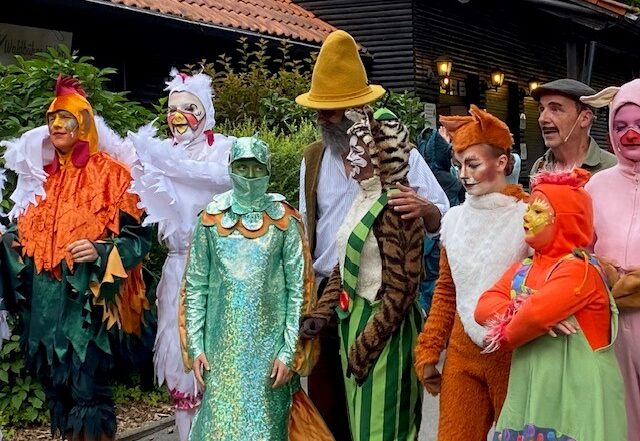 Eine Gruppe von Menschen in farbenfrohen Tier- und Figurenkostümen - darunter ein Huhn, eine Katze, ein Fuchs und eine Person mit grünen Schuppen - posieren im Freien auf der Waldbühne Heldritt und erwecken den Märchenzauber mit der Hochschule Coburg vor einem Holzgebäude und üppigem Grün zum Leben. at Hochschule Coburg