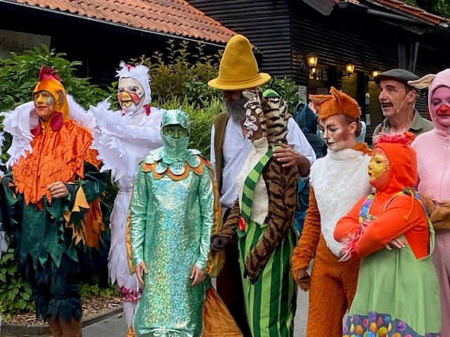 Eine Gruppe von Menschen in farbenfrohen Tier- und Figurenkostümen - darunter ein Huhn, eine Katze, ein Fuchs und eine Person mit grünen Schuppen - posieren im Freien auf der Waldbühne Heldritt und erwecken den Märchenzauber mit der Hochschule Coburg vor einem Holzgebäude und üppigem Grün zum Leben. at Hochschule Coburg