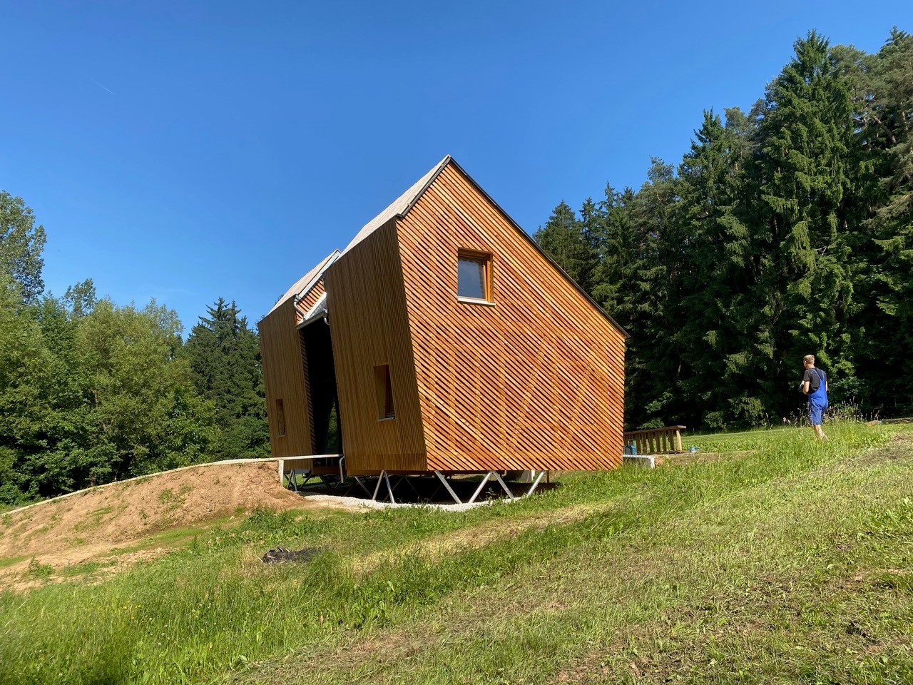 Auf einem grasbewachsenen Hang in der Nähe eines Waldes steht eine kleine, moderne Holzhütte auf Stelzen, gebaut mit einem nachhaltigen Modulsystem, und eine Frau in blauen Shorts steht in der Nähe unter einem klaren blauen Himmel. at Hochschule Coburg